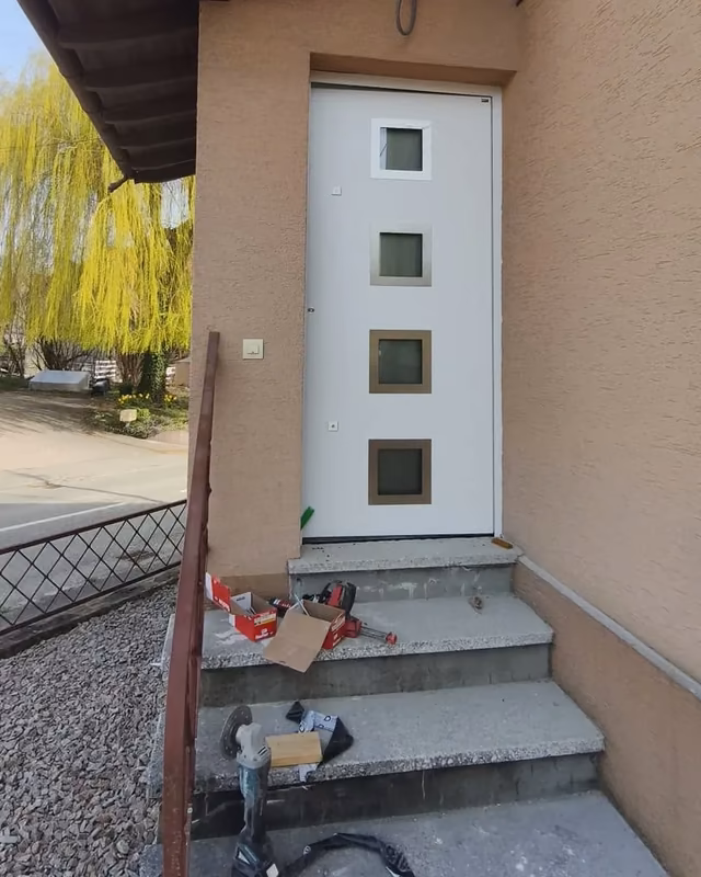 Porte d’entrée blanche à quatre petites fenêtres carrées, le jour de la pose, outils encore sur les marches — photo de chantier
